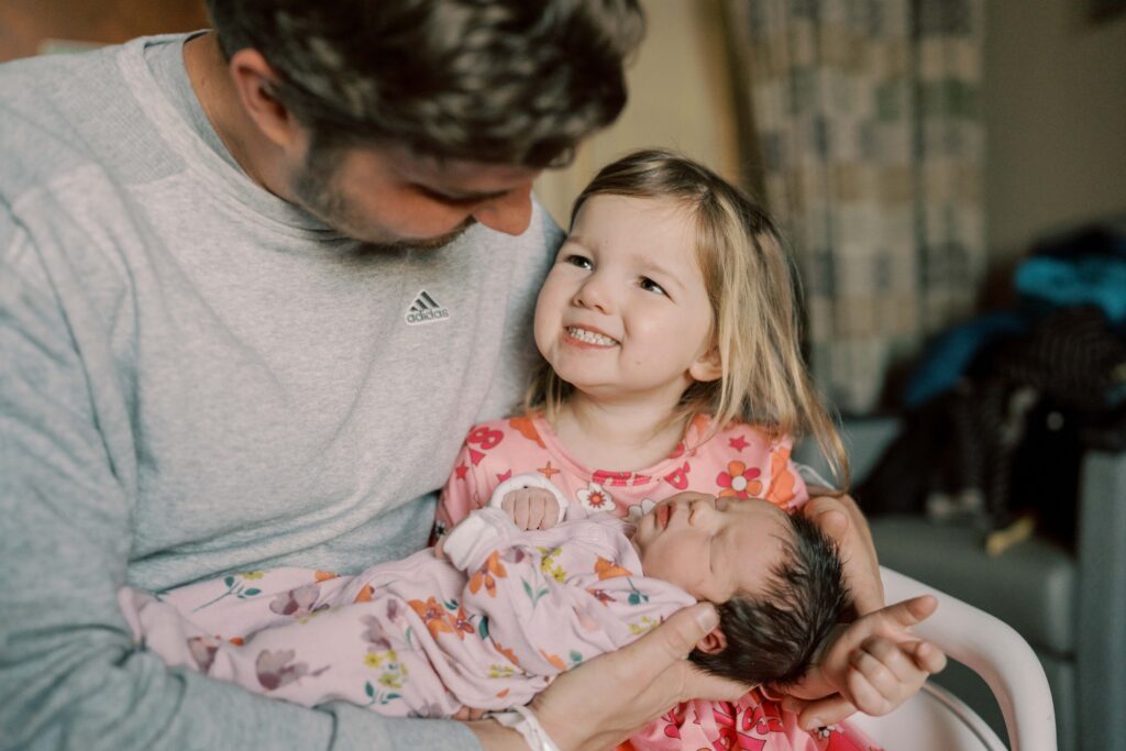 big sister giving dad a giant smile while holding her newborn baby sister
