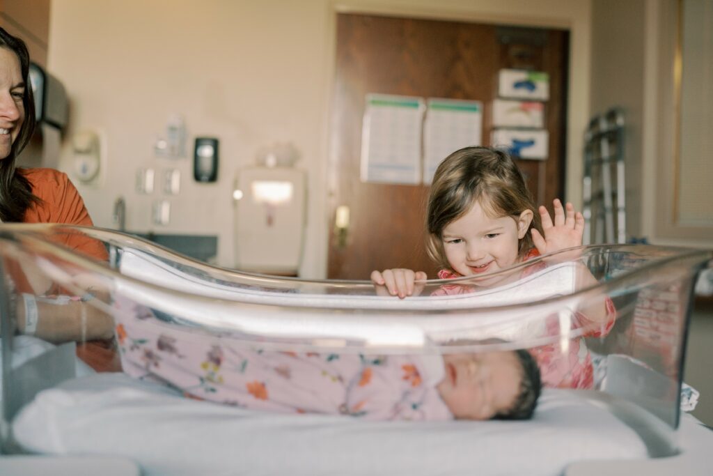 big sister waving at newborn baby in hospital bassinet during fresh 48 photos and film session